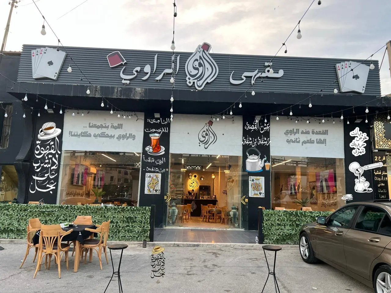 واجهة مقهى سوداء بحروف عربية بيضاء بارزة وأوراق لعب مجسّمة، مع ستيكر زجاج يحمل عبارات وخطوطًا عربية على كامل الفترينا.