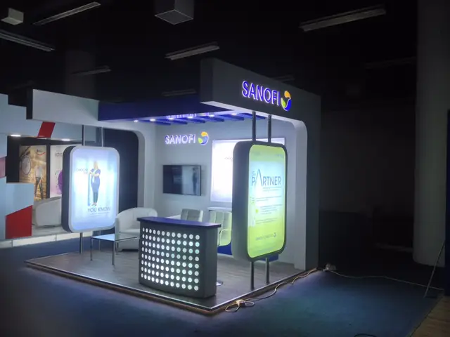 بوث معرض بواجهة علوية تحمل حروفًا مضيئة، ولوحات لايت بوكس رأسية، وكاونتر مثقّب مضيء، وجلسة ضيوف وشاشة عرض.
