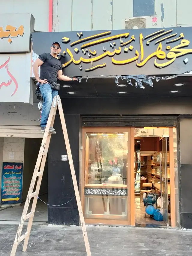 فنّي يقف على سلّم أمام واجهة محل مجوهرات أثناء تركيب حروف عربية ذهبية بارزة على لوحة سوداء.