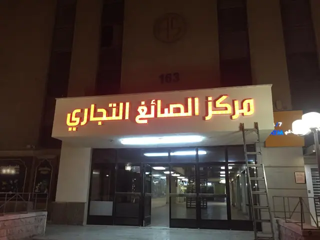 اسم المركز التجاري بحروف عربية مضيئة صفراء بحافة حمراء فوق مدخل المجمّع، مصوّرة ليلًا بعد التركيب.