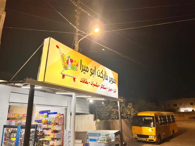 قارمة سوبرماركت صفراء مضاءة من الداخل تظهر ليلًا فوق واجهة المحل، مثبّتة على هيكل معدني بارز عن المبنى.