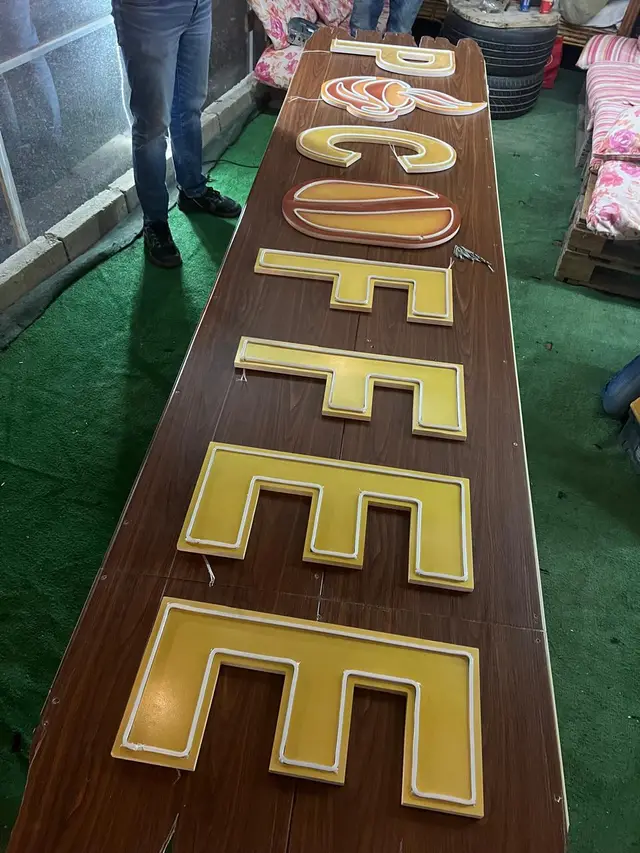 حروف كلمة COFFEE بوجه أكريليك أصفر وحافة نيون مرن مضيء، مصفوفة على لوح خشبي داخل الورشة قبل التركيب.