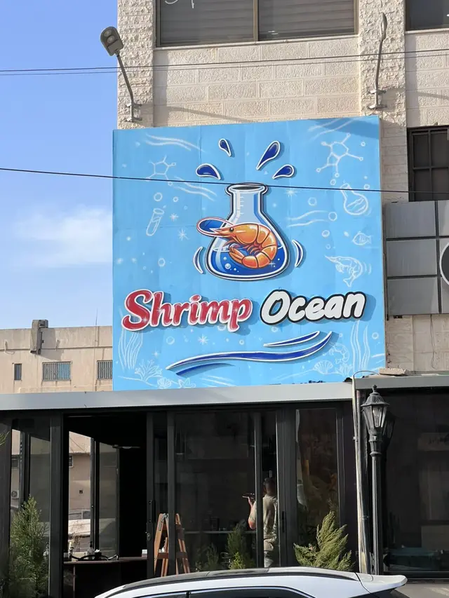 لوحة واجهة كبيرة مطبوعة بخلفية زرقاء ورسم دورق يحوي روبيان، مثبّتة فوق واجهة زجاجية لمطعم مأكولات بحرية.