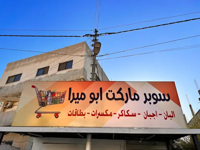 قارمة أفقية مطبوعة بتدرّج برتقالي تحمل اسم سوبر ماركت ورسم عربة تسوّق، مثبّتة على هيكل معدني فوق واجهة المحل نهارًا.