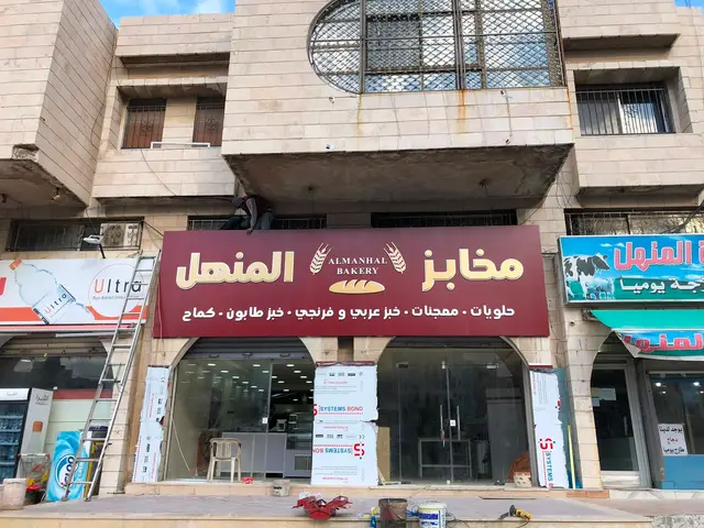 قارمة واجهة بعرض المحل كامل بخلفية عنابية وحروف بارزة بيضاء وذهبية وسطر خدمات أسفل الاسم، أثناء التركيب.